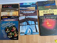 Photo showing a variety of vintage rock vinyl LP album covers arranged side by side on the floor including artists like Styx, Poppy Seeds, Neil Diamond, Queen, Creedence Clearwater Revival, and others.