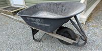 Side view of black metal wheelbarrow showing the basin, wooden handles, and no-puncture tire resting on gravel.