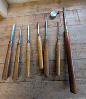 Seven wood turning chisels with wooden handles arranged on a wood surface.