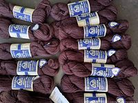 16 skeins of deep burgundy and purple-black Beehive French Tapestry Yarn wound in skeins with blue and white labels showing the brand and product details.