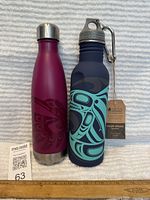 Two Indigenous Elements stainless steel water bottles on a white textured surface. One is purple with pink hummingbird design, the other is dark blue with light blue killer whale design and attached tag and clip.