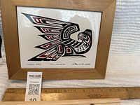 Photo of framed artwork with Thunderbird design, signed Clarence Wells 1982, depicting a stylized bird in black and red colors with wooden frame and ruler for scale.