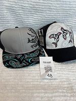 Photo showing two new adjustable baseball hats with Northwest Indigenous art. Left hat: gray and black with a Raven Fin Killer Whale graphic and turquoise accents on the bill. Right hat: white front panel with three stylized Killer Whales in black, red, and white on a black bill and back.