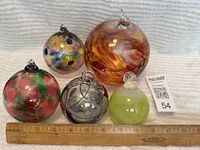 Five glass blown balls with distinct color patterns and hanging loops arranged alongside a ruler for size reference, measuring about 3 to 6 inches in diameter.