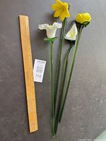 Four glass flowers with green stems next to a wooden ruler, showing approx 16 inches length