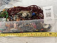 Plastic container filled with mixed broken jewelry pieces, beads, and craft items with a measuring tape showing about 30cm long.