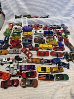 Overview of assorted Hot Wheels cars on a white cloth, showing various colors and styles including muscle cars, custom hot rods with exposed engines, sports cars, and trucks.