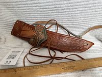 Full view of the leather sheath with straps, showing embossed floral patterns and attached leather laces, alongside a wooden ruler for size reference.