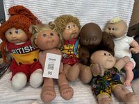 Lot of six vintage Cabbage Patch dolls arranged seated on a textured white surface showing diversity in hair colors and outfits.