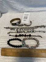 Top down photo of seven bracelets laid out on a white textured fabric with ruler for scale. Includes brass bangle, metal cuff, charm bracelet, bead bracelets.