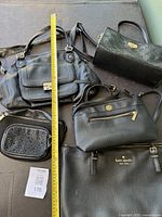 Overview of all 5 designer purses placed together on a table with measuring tape for scale, showing different styles and brands including Kate Spade and Anne Klein