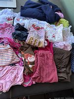Various infant girls clothing items laid out including shirts, pants, skirts, and jackets in assorted colors and prints, preowned condition.