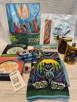 Mixed lot laid out showing calendar, plates, mugs, tea towel, cosmetic bag, and small carved stone and decorative rock with hand symbol.