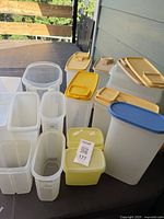 Overall view of assorted Tupperware containers of various sizes with mostly yellow, tan, and one blue lid on dark surface outdoors.