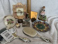 Photo showing entire lot including vanity items, decorative bowls, mirror, hair brush, trinket boxes, figurine, and embellishment piece on a soft textured background.