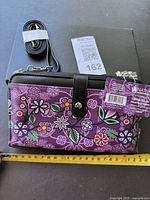 Front view of purple floral crossbody purse with black trim, measuring approximately 9 inches wide, accompanied by a rolled black strap and tag attached.