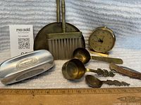 Vintage lot items grouped together on cloth background: a silver metal table brush, brass hand broom and dustpan, brass collector spoons, wooden handled knife, small brass clock.