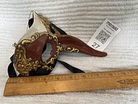 Full view of Wayne M Kleski Mardi Gras mask showing long beak nose with gold embellishments, brown and white colors, with ruler for size comparison.