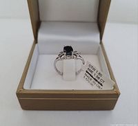 Ring displayed in gold-tone box showing front view with black oval stone and white gold filigree