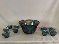 Photo showing the full set: one large carnival glass punch bowl and nine matching small cups arranged around it on a white cloth background.