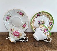 A pair of porcelain teacups and saucers featuring different floral designs and gold accents shown resting on a wooden surface against white background.