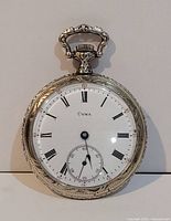 Front view of Cyma pocket watch showing white dial with Roman numerals, seconds sub-dial at six o'clock, and ornate engraved silver-tone case.