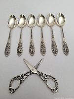 Set of six silver spoons laid out in a row with ornate handles and a pair of silver scissors below them showing the decorative scroll design.