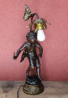 Full front view of the bronze lamp with child figurine and floral lamp holder, bulb installed, showing base and power cord.