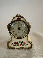 Full frontal photo of the German Reuge alarm clock in white porcelain with floral painting and gold decorative accents.