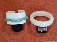 Two jade bracelets displayed on stands on a textured surface, showing both the white solid bangle and the green bead bracelet.