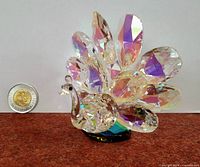 Full view of the crystal peacock figurine showing iridescent faceted tail feathers and black base alongside a coin for scale.