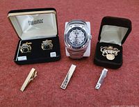 Full lot photo showing men's jewelry including wristwatch, cufflinks, tie clips, and lapel pin on reddish surface