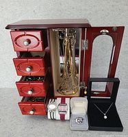 Full view of red wooden jewelry box with open drawers and mirrored side door exposing hanging necklaces, wristwatches, ring, pendant necklace, and earrings.