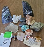 Collection of rocks and minerals including Orthoceras fossil bookends, quartz cluster, jasper, aurichalcite, chert, and basalt stones placed on a table with auction lot tag visible.