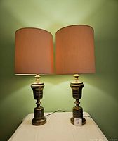 Pair of modernist brass table lamps on a white surface with beige shades and green background wall, showing size and condition of both lamps
