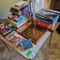 Folding card table with stacked vintage board games in boxes, including Mastermind, Catan, and Spot Cash, plus playing cards and a wooden dice shaker box.