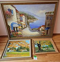 Three framed acrylic paintings displayed on the floor against a wall. Largest depicts a Mediterranean coastal scene with buildings, sea, and sky. Two smaller paintings show autumn scenes with trees and animals in pasture or ranch settings.