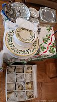 Lenox Holiday dinnerware collection displayed on holly-patterned cloth showing plates, teacups, silver-plated items, and chrome tray.