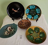 Five MCM decorative plates arranged on white fabric, showing various artistic enamel designs including roadrunner, horse, and floral motifs.