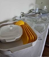 Vintage set including white plastic Kromex lazy susan serving tray with yellow corn holders, white casserole dish with lid, various glass serving trays, and silver-tone tray visible on table.