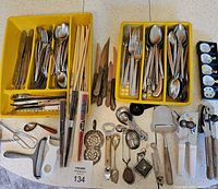 Two yellow trays with divided compartments holding mixed silverware including forks, spoons, knives, and sets of chopsticks. Various kitchen tools such as can openers, tea strainers, cheese slicers, peelers, ladles, and other utensils are arranged around the trays on a white speckled surface.