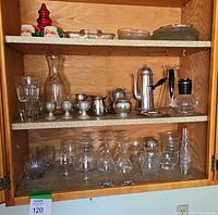 Cabinet shelf with a mix of clear glassware including glasses, goblets, a carafe, silver-tone teapot and cream/sugar set, ceramic Santa mugs, and a glass Christmas tree figure