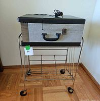 Full view of vintage Triomatic record player case on metal stand with wheels and record holder rack below.