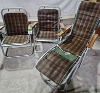 Front view of two vintage camp chairs and one camp lounger, all with matching green, orange, and brown plaid fabric and aluminum frames with wooden armrests. Lounger positioned reclined.