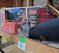 Multiple items displayed on shelf including boxed Coleman stove, metal firewood holder, Aladdin lamp with glass chimney, red plaid picnic bag, and red Autovox radio.