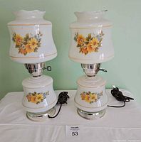 Two vintage milk glass hurricane lamps with yellow floral patterns and chrome bases seen from the front, cords coiled beside them.