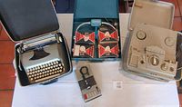 Photo of Smith Corona typewriter in black carrying case on left, magnetic tape recorder with tape in center, and beige Realistic tape recorder on right