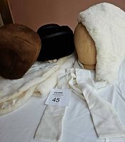 Three hats and a faux fur cape displayed on a surface with a wood hat form