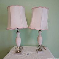Pair of vintage satin glass table lamps with white satin glass diamond-cut patterned bodies, square marble bases, and pale pink scalloped fabric lampshades showing tears inside.