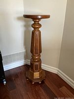 Side angle showing full height, round top, carved column, and octagonal base of the solid wood pedestal on hardwood floor near white baseboard.